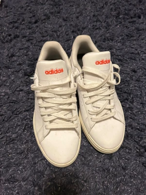 Vita sneakers från Adidas med röd logga - Fräscha vita sneakers från Adidas med röd logga på plösen. Skorna har snörning, rund tå och platt sula. Ovandelen är i syntetmaterial och har diskreta perforeringar på sidan för ventilation. Perfekt för dig som gillar klassisk stil med en modern touch.