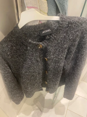 Grå fluffig kofta från Monki - Säljer en mysig grå kofta från Monki i storlek M. Koftan är fluffig och har en stickad känsla med snygga knappar framtill stil.