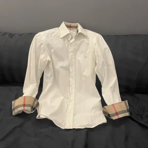 Vit Burberry skjorta slim fit XS - Snygg vit skjorta från Burberry i slim fit med klassiskt rutig mönster på insidan av krage och manschetter. Broderad Burberry-logga på bröstet. Skjortan har knappar framtill och är tillverkad i mjuk bomull med lite stretch för extra komfort.