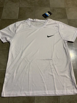 Vit Nike Dri-Fit t-shirt - Fräsch vit t-shirt från Nike med svart swoosh-logga på bröstet. Tillverkad i lätt och ventilerande Dri-Fit polyester som håller dig torr under träningen. Klassisk rund hals och korta ärmar, perfekt för sport och aktiva dagar.