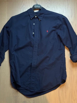 Marinblå skjorta från Polo Ralph Lauren - Säljer en marinblå skjorta från Polo Ralph Lauren i slim fit-modell. Skjortan har klassisk krage, knappar framtill och röd broderad logga på bröstet. Tillverkad i bomull med stretch för extra komfort. Perfekt för dig som gillar stilrena och snygga plagg.