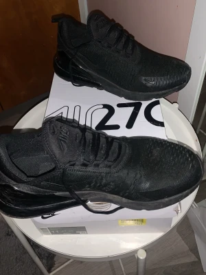 Nike Air Max 270 helsvarta sneakers - Nike Air Max 270 sneakers i helsvart färg med stickad ovandel och synlig Air-enhet i hälen. Skorna har snörning och platt sula, samt diskreta Nike-loggor på sidan och tungan. Perfekt för dig som gillar stilrena och sportiga sneakers.