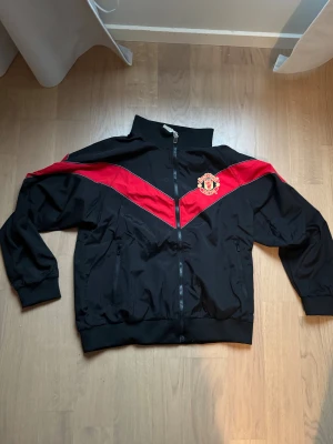 Manchester United svart och röd jacka - Säljer en svart Manchester United jacka med röda och vita detaljer i ett V-mönster över bröst och rygg. Jackan har klubbens emblem på framsidan och texten 'Manchester United 1878' broderad på ryggen. Hög krage, dragkedja och fickor med zip. Perfekt för fans!