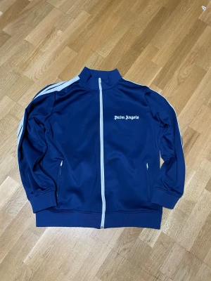 palm Angels  - Säljer en snygg marinblå zip-väst från Palm Angels med vita detaljer längs dragkedjan och axlarna. Västen har hög krage, två fickor med dragkedja och logga på bröstet. Tillverkad i mjuk polyester som ger en sportig vibe.