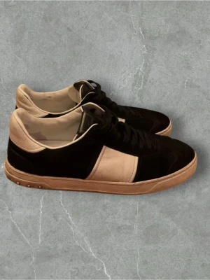 Valentino flycrew - Snygga Valentino Garavani sneakers i svart och beige med coola nitar på sulan. Skorna har rund tå, snörning och en platt sula. Materialet är en mix av skinn och mocka, vilket ger en lyxig känsla. Perfekt för dig som vill sticka ut med designerstil.