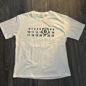 Vit t-shirt Maison Margiela med tryck - Snygg vit t-shirt från Maison Margiela med det ikoniska siffertrycket och en markerad 6:a på bröstet. Klassisk passform, rund hals och korta ärmar. Materialet känns väldigt exklusivt och är skön mot huden, Endast använd 1 gång med tagg.