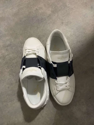 Valentino Garavani Open sneakers vit/blå - Säljer ett par snygga Valentino Garavani Open sneakers i vitt skinn med bred mörkblå läderdetalj över snörningen. Skorna har rund tå, platt sula och klassisk logga på plösen. Perfekt för dig som gillar exklusiva och stilrena sneakers. Priset kan diskuteras vid snabb affär!