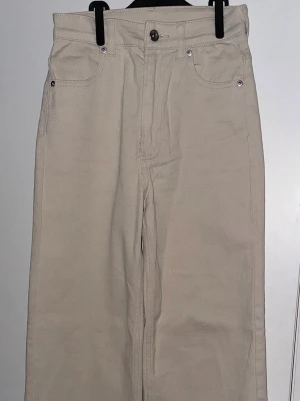 Beige raka jeans från H&M  - Snygga beige jeans från H&M Divided i rak modell med fem fickor och knappstängning. Byxorna har hög midja och är tillverkade i ett slitstarkt bomullsmaterial. Perfekt för dig som gillar en clean och enkel stil.