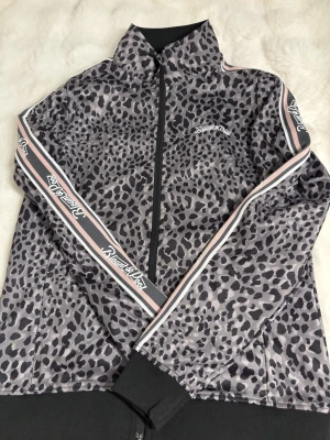 Leopardmönstrad vindjacka Blount & Pool - Snygg vindjacka med leopardmönster från Blount & Pool. Jackan har svarta och grå toner, dragkedja framtill och coola band med logga längs ärmarna. Perfekt för dig som vill sticka ut med streetstyle och sportig vibe.