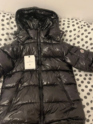Svart Moncler pufferjacka med huva - Säljer en svart pufferjacka från Moncler med glansig finish och huva. Jackan har dragkedja framtill, två sidofickor med dragkedja och Moncler-logga på ärmen. Insidan har en cool serietidningspatch. Perfekt för kalla dagar!