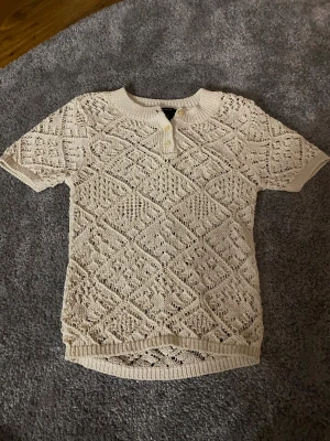 Beige stickad kortärmad tröja Lindex - Säljer en beige stickad tröja från Lindex i storlek XS. Tröjan har korta ärmar, rund hals och dekorativa knappar framtill. Det öppna stickade mönstret ger en cool och luftig känsla, perfekt för varmare dagar. Passar dig som gillar detaljer och unik stil.