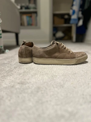 Bruna Lanvin sneakers i mocka - Säljer ett par bruna sneakers från Lanvin i mjuk mocka med rund tå och platt sula. Skorna har snörning och en beige yttersula med diskret logga undertill. Perfekta för dig som gillar stilrena och klassiska sneakers med lyxig känsla.