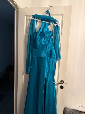 Blå långklänning från Anny Lee - Snygg blå långklänning från Anny Lee i glansig polyester med 5% spandex. Klänningen har halterneck, markerad midja med veckade detaljer och medföljande sjal. Passar perfekt för speciella tillfällen och har en elegant slits framtill.