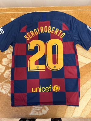 Barça fotbollströja Sergi Roberto #20 - Barcelona matchtröja med rutigt mönster i marinblått och vinrött, tryck med Sergi Roberto och nummer 20 i gult på ryggen. La Liga-patch på ärmen, Rakuten och Beko-loggor samt klubbmärke på bröstet. Tillverkad i polyester, perfekt för fotbollsfans.