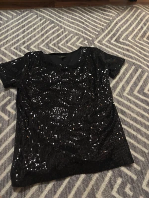 Svart paljett topp från Carla F - Säljer en svart topp från Carla F som är täckt med glittriga paljetter. Toppen har rund halsringning och korta ärmar, perfekt för att sticka ut med lite extra bling. Grym att styla med jeans eller kjol