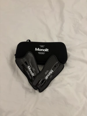 Monolit benskydd i kolfiber - Säljer ett par svarta benskydd från Monolit i kolfiber med vit logga. De är lätta och smidiga, perfekta för dig som vill ha maxad rörelsefrihet på planen. Kommer med ett praktiskt svart fodral för enkel förvaring.