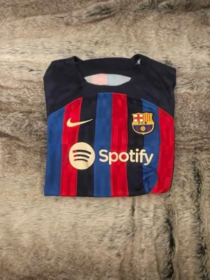 FC Barcelona Pedri matchtröja Nike - Säljer en officiell FC Barcelona matchtröja från Nike med Pedri och nummer 8 på ryggen. Tröjan har klassiska blå och röda ränder, klubbmärke och Nike-logga i guld samt Spotify som sponsor framtill. Perfekt för dig som älskar Barca och vill ha en snygg supportertröja.