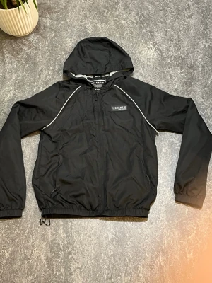 Svart vindjacka från McKenzie, S - Svart vindjacka från McKenzie med huva och grå meshfoder. Jackan har vita detaljer längs axlarna, dragkedja framtill och elastiska muddar vid ärmslut och nederkant. Perfekt för blåsiga dagar och har sportig vibe med logga på bröstet.