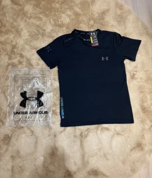 Svart Under Armour t-shirt i polyester - Svart t-shirt från Under Armour med korta ärmar och rund hals. Materialet är mjukt och stretchigt polyester, perfekt för träning. Diskret logga på bröstet och färgglad Under Armour-text längs sidan. Snygg och sportig look för dig som gillar att hålla igång. Kom dm för eventuella frågor 🧐 pris kan diskuteras 💯 T-shirt kommer med sin förpackning 🤩har aldrig används är i nytt skick. 