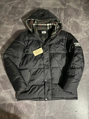 Svart pufferjacka från Burberry  - Svart pufferjacka från Burberry med avtagbara ärmar så du kan bära den som väst. Jackan har huva med rutigt foder, dragkedja och knappar framtill samt Burberry-logga på ärmen. Perfekt för kalla dagar och riktigt snygg streetstil.