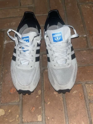 Adidas La trainer - Säljer ett par snygga Adidas sneakers med klassiska svarta ränder på sidorna och blå logga på plösen. Skorna är grå med mesh och mockadetaljer, har snörning och rund tå. Perfekta för dig som gillar retrostil och sportig vibe.