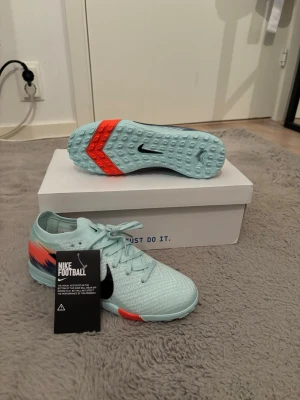 Nike Zoom Flyknit fotbollsskor ljusblå Barn - Nike Zoom Flyknit fotbollsskor i ljusblå färg med svarta swoosh-loggor och coola detaljer i orange och mörkblått på hälen. Skorna har snörning, platt sula med mönstrad undersida och är tillverkade i stickad syntet för grym komfort och lätt känsla på planen.