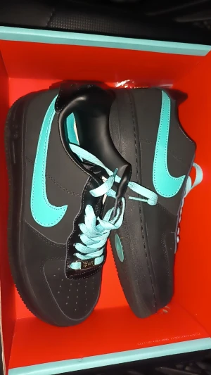 Nike Air Force 1 svart/turkos - Säljer ett par Nike Air Force 1 sneakers i svart med turkosa detaljer och snörning. Skorna har en platt sula och rund tå, med klassisk Nike-logga i turkos på sidan. Materialet är skinn och syntet, och de kommer i originalkartong.