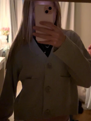 Beige kofta med stora knappar - Säljer en beige kofta med v-ringning och tre stora knappar framtill. Koftan har två fickor och ribbade muddar vid ärmslut och nederkant. Modellen är croppad och har en loose passform, perfekt att slänga på över en t-shirt eller linne.