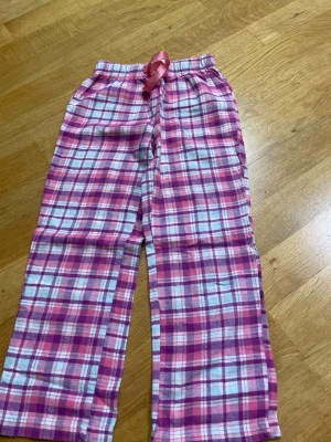 Rosa-rutiga mjukisbyxor från Lupilu - Säljer ett par mjuka och sköna mjukisbyxor från Lupilu i storlek 110/116. Byxorna har ett rutigt mönster i rosa, lila och vitt samt en bred resår i midjan med rosa snörning. Perfekta för chill hemma eller mysiga dagar.