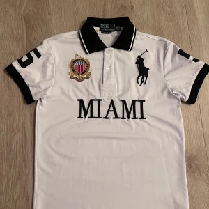 Vit polo från Ralph Lauren - Säljer en vit polo från Ralph Lauren med svart märke och svarta detaljer på ärmarna. Toppen har broderad logga, emblem och texten MIAMI framtill samt en stor svart siffra 5 på ryggen. Tillverkad i bomull och har klassisk pikédesign med knappar vid halsen.