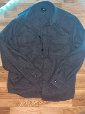 Mörkgrå overshirt från H&M - Säljer en stilren mörkgrå overshirt från H&M med klassisk krage och två stora bröstfickor med lock. Jackan har knappar framtill och vid ärmsluten, perfekt att slänga på över en hoodie eller t-shirt. Materialet är mjukt och känns skönt mot huden.