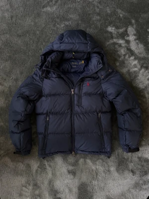Mörkblå pufferjacka från Polo Ralph Lauren - Mörkblå pufferjacka från Polo Ralph Lauren med huva och dragkedjefickor. Skön och tjock jacka med klassisk röd Polo-logga, perfekt för vardagen och kalla dagar.