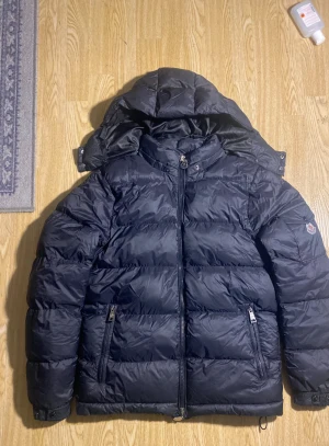 Svart Moncler pufferjacka med huva - Svart pufferjacka från Moncler med avtagbar huva, två dragkedjefickor och coolt serietryck på insidan. Jackan har en quiltad design och justerbar dragsko nedtill. Perfekt för dig som vill ha en varm och snygg vinterjacka med exklusiv känsla.