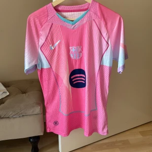 Barça x Nike rosa fotbollströja M - Säljer en oanvänd Barcelona fotbollströja från Nike i rosa med ljusblå och lila detaljer. Tröjan har korta ärmar, V-ringad krage med turkos kant och coola grafiska linjer framtill. Materialet är lätt och andas, perfekt för träning eller match. Spotify-logga på bröstet.