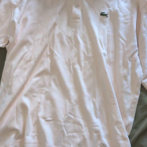 Vit piké från Lacoste med krage - Snygg vit pikétröja från Lacoste med klassisk krage och två knappar framtill. Den har det ikoniska gröna krokodilmärket broderat på bröstet. Tillverkad i mjuk bomull som känns skön mot huden. Perfekt för dig som gillar stilren och enkel design. Strl L