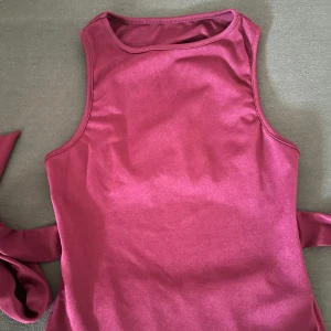 Vinröd omlottopp med knytband - Säljer en vinröd ärmlös omlottopp med breda knytband i midjan och öppen rygg. Toppen har en snygg cut-out detalj baktill och är tillverkad i ett mjukt, stretchigt material. Perfekt för dig som vill ha en trendig och unik look.