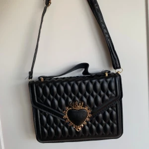 Svart quiltad axelväska med hjärtdetalj - Quiltad svart axelväska i syntet med justerbar rem och handtag. Väskan har en unik guldig hjärtdetalj framtill med krona och dekorativa inslag. Stilren och edgy design som sticker ut, perfekt för dig som gillar statement-accessoarer.
