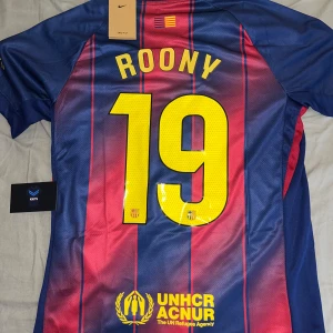 Barcelona matchtröja 25/26 Roony - Snygg FC Barcelona matchtröja från Nike med Roony och nummer 19 på ryggen. Tröjan har klassiska blå och röda vertikala ränder, gula detaljer och klubbmärke på bröstet. Spotify-logga och UNHCR-tryck. Tillverkad i lätt, ventilerande polyester med korta ärmar.
