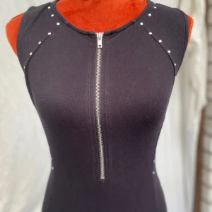 Svart kort klänning från H&M Divided 38 - Svart, ärmlös klänning med dragkedja framtill och dekorativa nitar längs axlarna och sidorna. Figurnära passform. ﻿95% bomull, 5% elastan Längd: 82 cm ﻿Bystvidd: 43 cm  Inga bilder när den sitter på mig! Katter finns i hemmet! 01