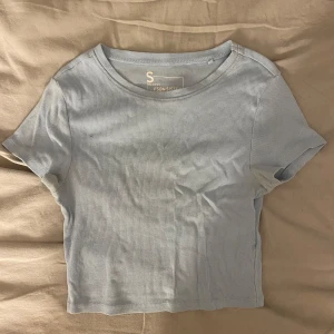 Ljusblå ribbad topp från new yorker - En ljusblå croppad topp från new yorker i ribbad bomull. T-shirten har korta ärmar och rund halsringning, perfekt för en chill och enkel stil. Snygg att matcha med jeans eller kjol för en avslappnad vibe.