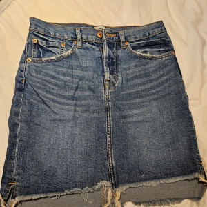 Blå jeanskjol med rå kant från 157 - Snygg blå jeanskjol från 157 i storlek S. Kjolen har klassiska fickor fram och bak, knapp och dragkedja i midjan samt en rå, fransig kant nedtill som ger en cool vibe. Perfekt för dig som gillar denim och vill ha en kortkjol med edge.