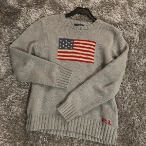Grå stickad tröja Polo Ralph Lauren - Snygg grå stickad tröja från Polo Ralph Lauren med amerikansk flagga på bröstet och RL-broderi nertill. Tröjan har rund halsringning, långa ärmar och breda ribbade muddar. Perfekt för dig som gillar klassisk och stilren design.