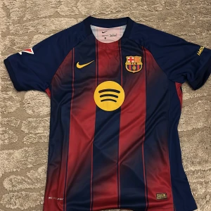 FC Barcelona Lamine Yamal tröja M - Säljer en FC Barcelona matchtröja med Lamine Yamal och nummer 10 på ryggen. Tröjan är kortärmad, har klassiska blå och röda vertikala ränder, gula detaljer och sponsorloggor. Tillverkad i Dri-FIT material som andas, perfekt för fotboll eller häng med polarna. Köpte i Barcelona.