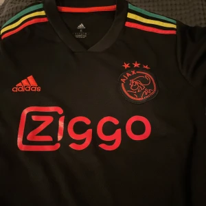 Ajax svart kortärmad fotbollströja Adidas - Ajax svart fotbollströja från Adidas med röd Ziggo-logga och klubbmärke på bröstet. Färgglada ränder i rött, gult och grönt på axlarna ger en unik vibe. Tröjan har V-ringad krage och är tillverkad i lätt, ventilerande material perfekt för match eller träning.