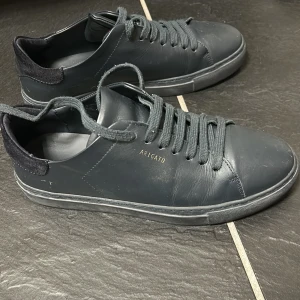 Svarta sneakers från Axel Arigato - Stilrena svarta sneakers från Axel Arigato med diskret guldfärgad logga på sidan. Skorna har ovandel i skinn, svart mockadetalj vid hälen och klassisk snörning. Perfekta för dig som gillar minimalistisk och clean stil. Pris kan även diskuteras.