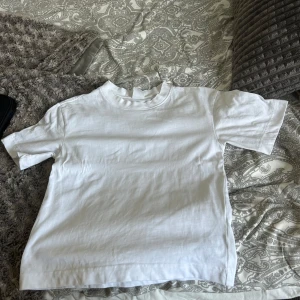 Vit basic t-shirt med rund hals - En clean och enkel vit t-shirt med rund hals och korta ärmar. Modellen är rak och har en normal passform, perfekt för att matcha med jeans eller shorts. Materialet känns mjukt och skönt mot huden, troligtvis bomull. Inga synliga loggor eller detaljer. Aldrig använt denna och den är som helt ny, säljer för den är alldeles för liten och kunde inte skicka tillbaka.