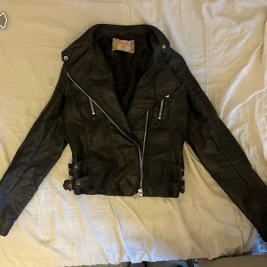 Svart skinnjacka från chiquelle - Snygg svart skinnjacka från Voyelles med coola detaljer som silvriga dragkedjor, bred krage och spännen i midjan. Jackan har lång ärm och en croppad passform. Storlek 38 men skulle säga att den passar både xs och s.