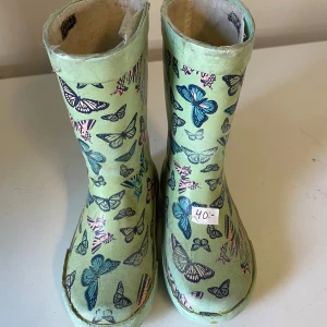 Gröna stövlar med fjärilsmönster - Snygga gröna stövlar med färgglada fjärilar i blått, rosa och svart. Perfekta för regniga dagar och ger en lekfull touch till outfiten. Hög modell med rund tå och platt sula.