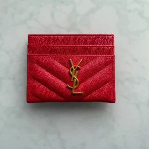 Röd plånbok från Saint Laurent - Säljer en snygg röd plånbok från Saint Laurent med guldigt YSL-emblem framtill. Plånboken är i skinn med quiltad struktur och har flera kortfack. Perfekt för dig som vill ha något stilrent och lyxigt i fickan.