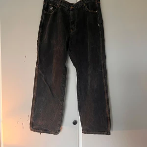 Svarta wide jeans med broderad logga - Säljer ett par svarta jeans med bred passform och slitna detaljer. Byxorna har orange kontrastsömmar, broderad logga på fickan och råa kanter vid bensluten. Materialet är klassisk denim och modellen har flera fickor, inklusive bakfickor med brodyr.